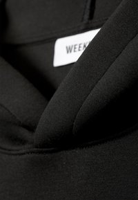 Črn oblačilo iz tkanine s prikazom bližnjega posnetka ravnega ovratnika. Material je videti gladek s finim teksturiranim vzorcem. Vidna je oznaka z napisom "WEEKDAY".