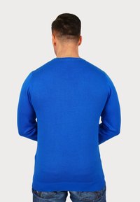 Modra pletena jopica z dolgimi rokavi, okroglo ovratnike in rebrastim robom, kombinirana s svetlo modrimi kavbojkami. Material se zdi gladek in prilega.