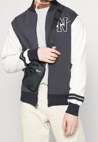 Donkergrijs-witte bomberjack met "N"-logo, geribbelde manchetten en ritssluiting. Gedragen over een grijs shirt met een zwarte Nike crossbodytas.