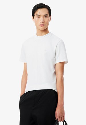 Junger Mann trägt ein schlichtes weißes T-Shirt mit kurzen Ärmeln und schwarze Hose, hält mit seiner rechten Hand einen schwarzen Taschengriff.