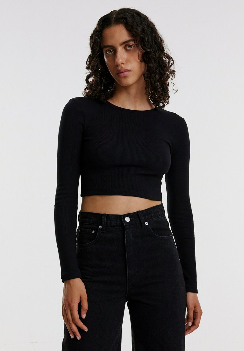 Schwarzes, geripptes Langarm-Crop-Top, kombiniert mit hochgeschnallten schwarzen Jeans. Es hat einen runden Halsausschnitt und eine figurbetonte Silhouette.