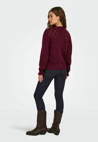 Pull en maille bourgogne avec motif en losange, poignets et ourlet côtelés, porté avec un jean noir et des bottes de cowboy marron, vu de dos.