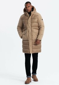 Beige Pufferjacke mit Kapuze, Druckknöpfen und zwei vorderen Taschen. Ein Paar schwarze Jeans und braune Wildleder-Stiefeletten. Glatte Textur, lässiges Design.