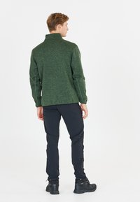 Grön, strukturerad zip-jacka med hög krage, bärs med svarta byxor och stövlar; analyserad från baksidan, vilket avslöjar jackans design.