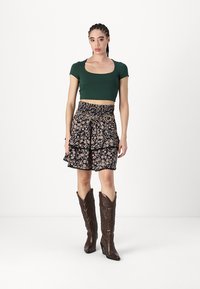 Zöld crop top, virágmintás fekete szoknya fodrokkal és csipkés díszítéssel, barna cowboy csizma texturált részletekkel, fehér háttér előtt áll.