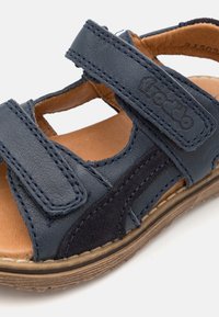 Mörkblå lädersandal med kardborreband, mockadetaljer, synlig sömnad och en brun innersula. Designad för komfort och andningsförmåga.
