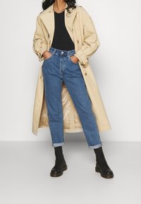 Un abrigo trench beige sobre una blusa negra, jeans azules remangados y botas negras con cordones. El abrigo presenta un diseño clásico con detalles de botones.