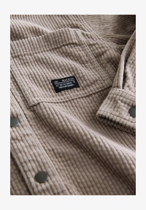 Bézs kordbársony anyag függőleges bordázattal, fekete címkés varrott folttal és ezüst gombokkal. Puha tapintású, klasszikus dizájn.