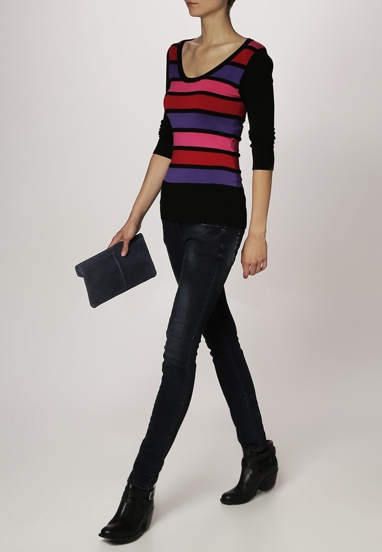 Pruhovaný svetr s černými, červenými, fialovými a růžovými vodorovnými pruhy; úzké modré džíny; drží šedé psaníčko; nosí černé kotníkové boty.