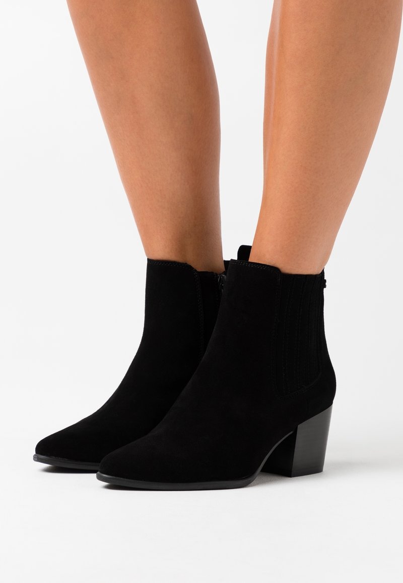 Czarne zamszowe botki za kostkę na słupku, noszone na nagich nogach, z elastycznymi panelami po bokach i zamkiem błyskawicznym z boku.