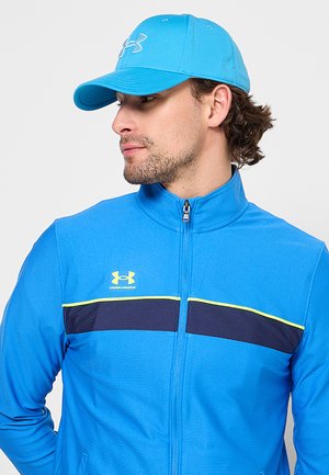 Homme portant une casquette bleu vif Under Armour et une veste zippée assortie avec une bande noire et jaune sur la poitrine.