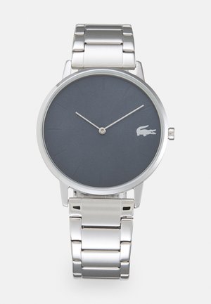 Montre en acier inoxydable argenté avec un cadran rond gris foncé. Présente des marqueurs d'heure minimalistes et un petit logo blanc sur le cadran.