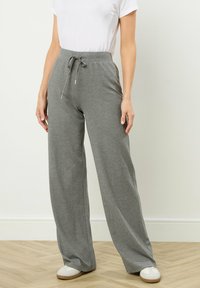 Vrouw in hoge, grijze, wijde broek met een koord, gecombineerd met witte sneakers en een kort mouw, wit shirt dat in de broek is gestopt.