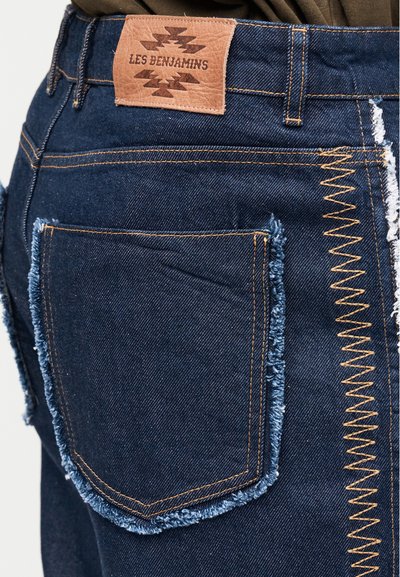 Σκούρο μπλε τζιν παντελόνι με ξεφτισμένες άκρες στις τσέπες και ζιγκ-ζαγκ ραφές, με ταμπέλα από δέρμα ταμπά με την επιγραφή "LES BENJAMINS" στη ζώνη της μέσης.
