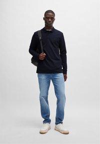 Maritieme langekorte polo, blauwe jeans en witte sneakers. Model houdt een zwarte tas vast en staat tegen een effen lichte achtergrond.