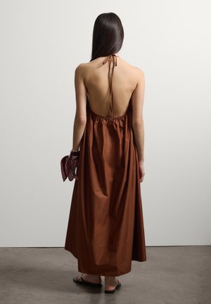 Mujer con cabello largo y oscuro, vestida con un vestido halter marrón sin espalda y sosteniendo una bufanda roja, de pie sobre un suelo de hormigón.