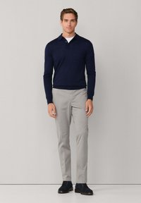 Granatowa koszula polo z długim rękawem i płaskim kołnierzykiem, zestawiona z jasnoszarymi spodniami i granatowymi zamszowymi butami, stojąca na tle w neutralnych kolorach.