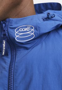 Blaue Kapuzenjacke aus glattem Stoff mit durchgehendem Reißverschluss, Kordelzugakzenten und gesticktem "Project Core"-Logo am Kragen.