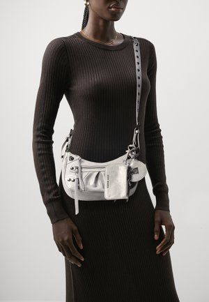 Vrouw in een zwart geribbeld jurkje met een zilveren schoudertas die meerdere ritszakken en bevestigde zakjes heeft.