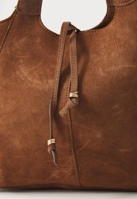 Sac fourre-tout en suédine marron avec une ouverture arrondie, des accents noués et des détails en métal doré, offrant une texture douce et un design simple.
