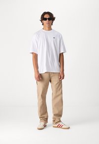 T-shirt bianco in cotone, pantaloni cargo beige e sneakers color crema con strisce rosse, indossati da un modello maschile con occhiali da sole scuri e capelli mossi.