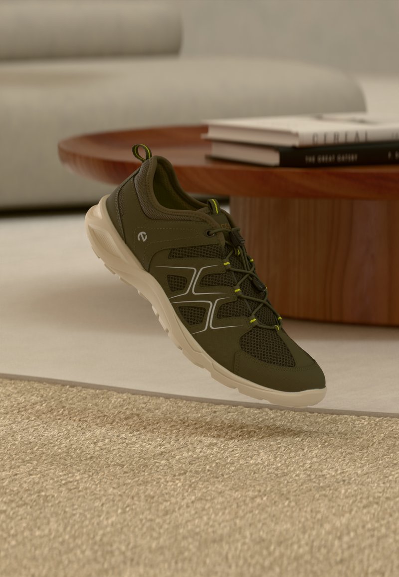 Baskets vert olive avec semelle beige, panneaux en mesh et lacets élastiques flottant au-dessus d'un tapis beige dans un salon moderne.