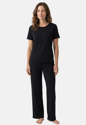 Kvinde med bølget brunt hår iført en ensfarvet sort kortærmet t-shirt og løse sorte bukser, stående barfodet mod en hvid baggrund.