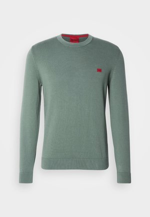 Pull en tricot vert à manches longues, col rond et poignets côtelés. Présente un petit logo rouge sur la poitrine. Texture lisse, design minimal.