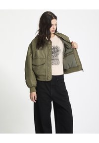 Oliven grøn bomberjakke med oversized pasform, ribbede manchetter og to forlommer, båret over en lyserød t-shirt med grafik og sorte bukser.