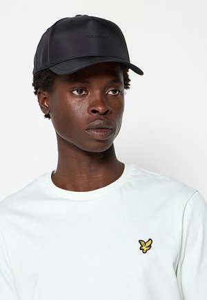 Ung person iført en sort kasket og hvid skjorte med en gul fuglelogo, der kigger let til højre på en ensfarvet lys baggrund.