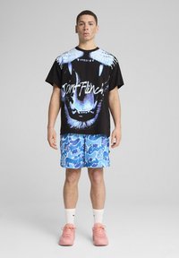 Sort t-shirt med en blå grafik af et bjørnes ansigt og teksten "Don't Flinch," sammen med blå shorts med abstrakte designs, båret med lyserøde sneakers.