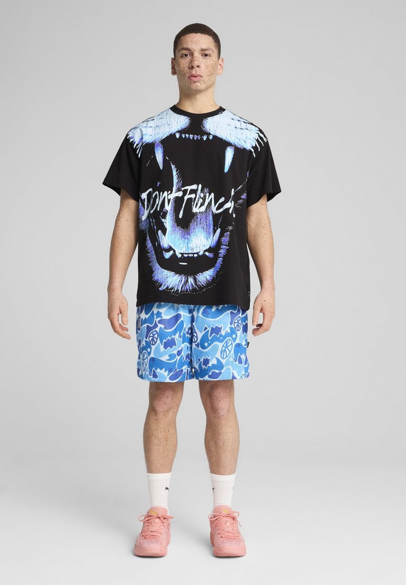 Svart t-shirt med en blå grafisk bild av en björnansikte och texten "Don't Flinch," tillsammans med blå shorts med abstrakta mönster, bärs med rosa sneakers.