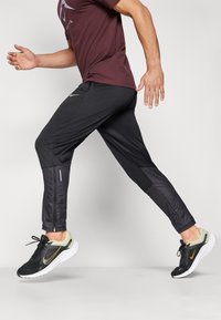 Calças atléticas pretas com tecido texturizado e painéis lisos, apresentando detalhes refletivos e um ajuste afunilado, combinadas com ténis pretos e amarelos.