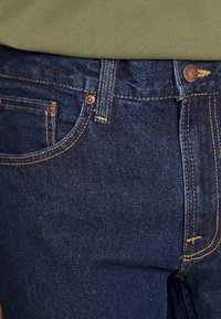 Tmavě modré džínové kalhoty s kováním v barvě mědi, viditelným prošíváním a klasickým designem s pěti kapsami, zobrazené s zeleným topem nahoře.