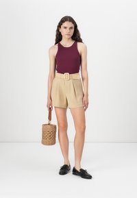Burgundy sleeveless top, beige high-waisted shorts with a woven belt, black shoes, and a round woven bag with a wooden handle.