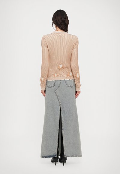 Femeie purtând un pulover bej deteriorat din tricot și o fustă lungă gri din denim cu o despicătură în spate, și pantofi negri cu toc, stând cu spatele la privitor.