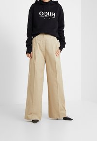 Felpa nera con cappuccio e logo bianco, abbinata a pantaloni beige a gamba larga; texture liscia, vestibilità sartoriale e scarpe nere appuntite e affusolate.