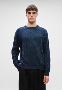 Marineblåstrikket genser med et strukturelt diamantmønster, rund hals og ribbestrikkede mansjetter og nederkant, kombinert med svarte bukser.