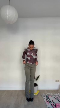 Kvinde med håret i en knold iført blomsterprintet top og olivengrønne bukser, stående i et minimalistisk rum med hvide vægge, trægulv, tæppe, plante og rund loftslampe.