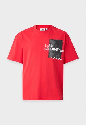 Мъж позира с яркочервена тениска Off-White с черна графика на гърдите и тъмен панталон на изчистен бял фон.