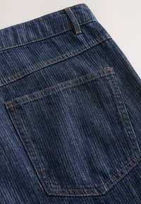 Jeans din denim de culoare albastră închisă, cu un buzunar din spate cu cusături portocalii. Țesătura are o textură subtilă în dungi și un design clasic.