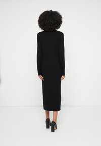 Fekete, hosszú ujjú, testhezálló ruha midi hosszúságban, sima anyagból, egyszerű, kerek nyakkivágással. Fekete magassarkú cipővel viselve.