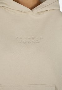 Felpa beige con tasca frontale, polsini a coste e logo in rilievo che recita "PEGADOR Messengers of Truth". Tessuto morbido.