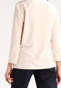 Donna che indossa un top beige a maniche lunghe e jeans blu scuri, vista da dietro su uno sfondo bianco.
