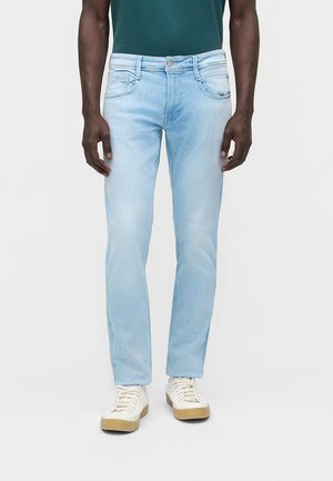 Mann trägt hellblaue Slim-Fit-Jeans, dunkelgrünes Hemd und weiße Turnschuhe und steht vor einem schlichten weißen Hintergrund.