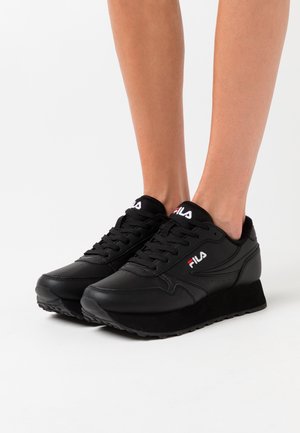 Person, die schwarze Fila Plateau-Sneaker mit Schnürdesign auf weißem Hintergrund trägt, zeigt Unterschenkel und Füße.