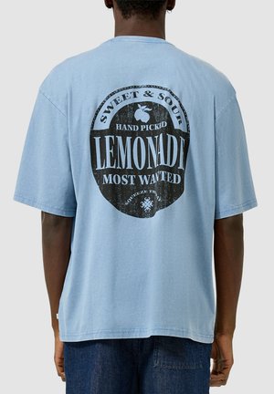 Mężczyzna noszący jasnoniebieski, oversize'owy t-shirt z czarną, okrągłą grafiką i napisem „Sweet & Sour Hand Pickd Lemonade Most Wanted” na plecach.