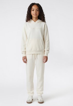 Giovane donna in piedi che indossa una felpa Champion color crema, pantaloni della tuta abbinati e sneakers bianche su uno sfondo semplice.