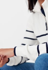 Persona che indossa un maglione bianco con strisce blu, seduta con le mani appoggiate sul ginocchio in jeans blu, contro uno sfondo bianco semplice.