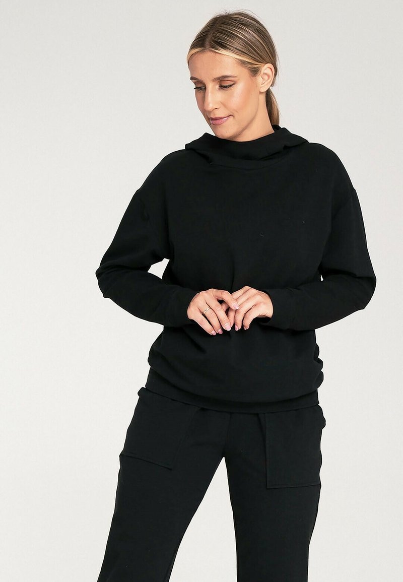 Czarny sweter z kapturem, z długimi rękawami i luźnym krojem. Wykonany z miękkiego materiału; noszony z pasującymi czarnymi spodniami z kieszeniami po bokach.